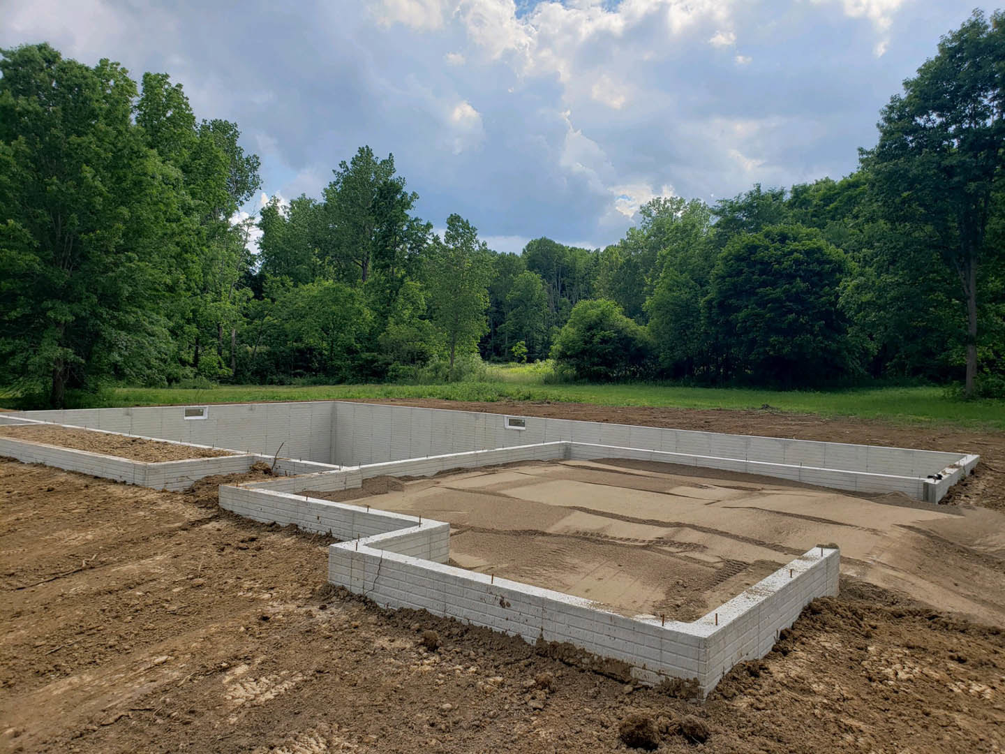Concrete house foundation with framed window opening, surrounded by soil and grass, under blue sky with scattered clouds and trees in background