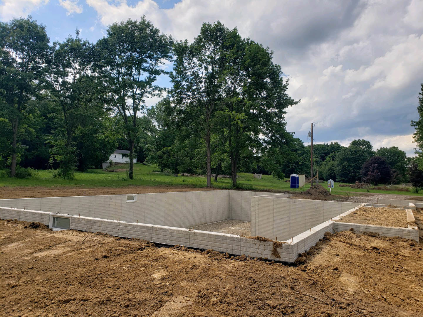 Grassy building site bordered by mature trees, dirt area in foreground, white structure with square window visible among foliage