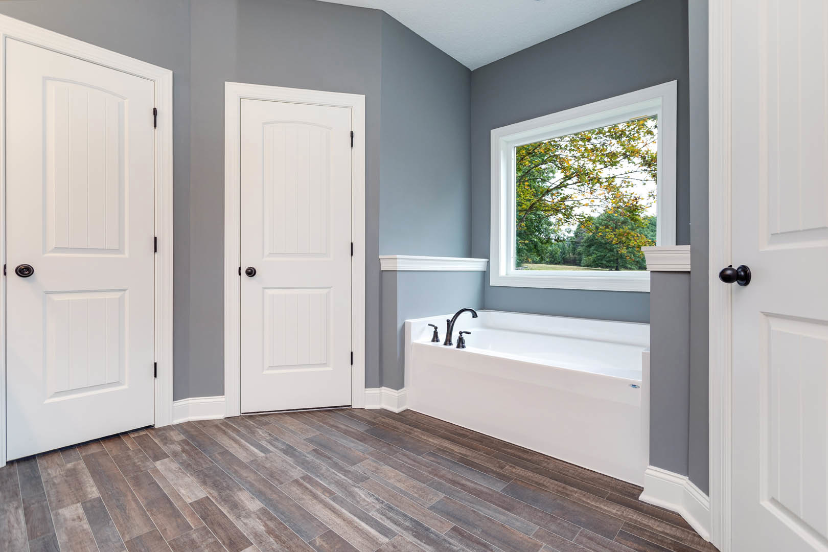 Bathroom featuring a freestanding white bathtub with chrome faucets, wood plank flooring, two white doors with black hardware, and a window overlooking trees.