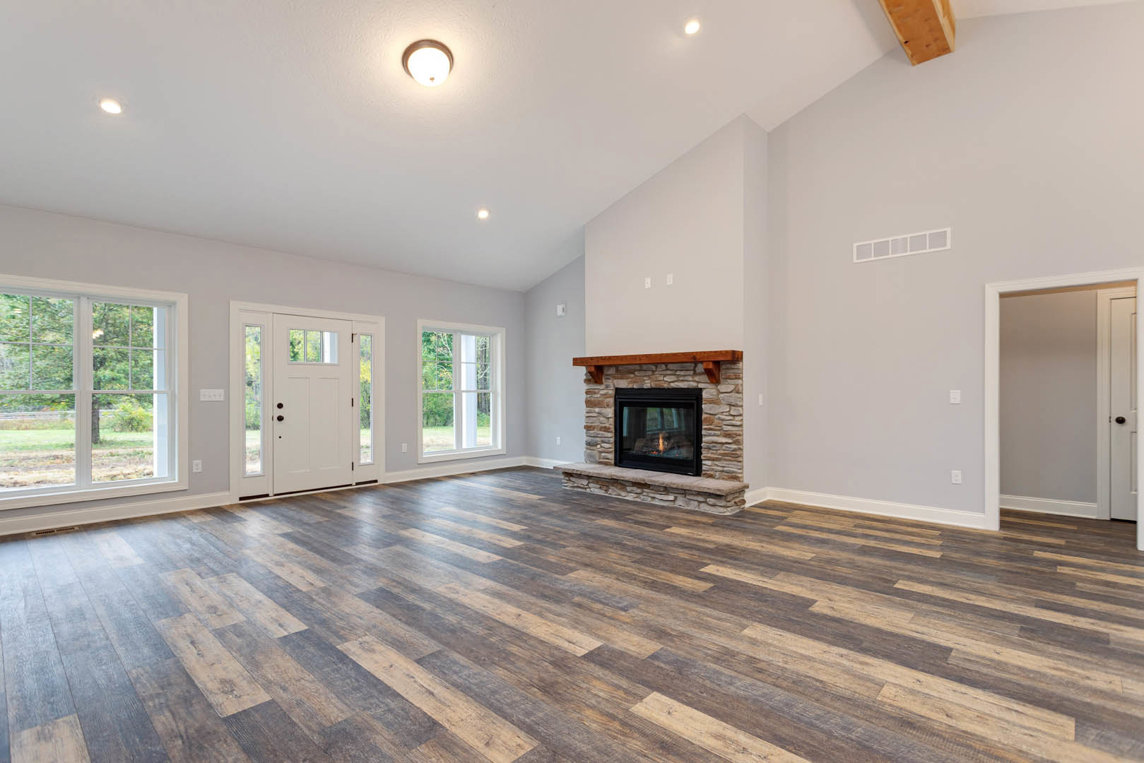 Living room with hardwood floors, stone fireplace with wooden mantel, ceiling light fixture, large window showing outdoor view