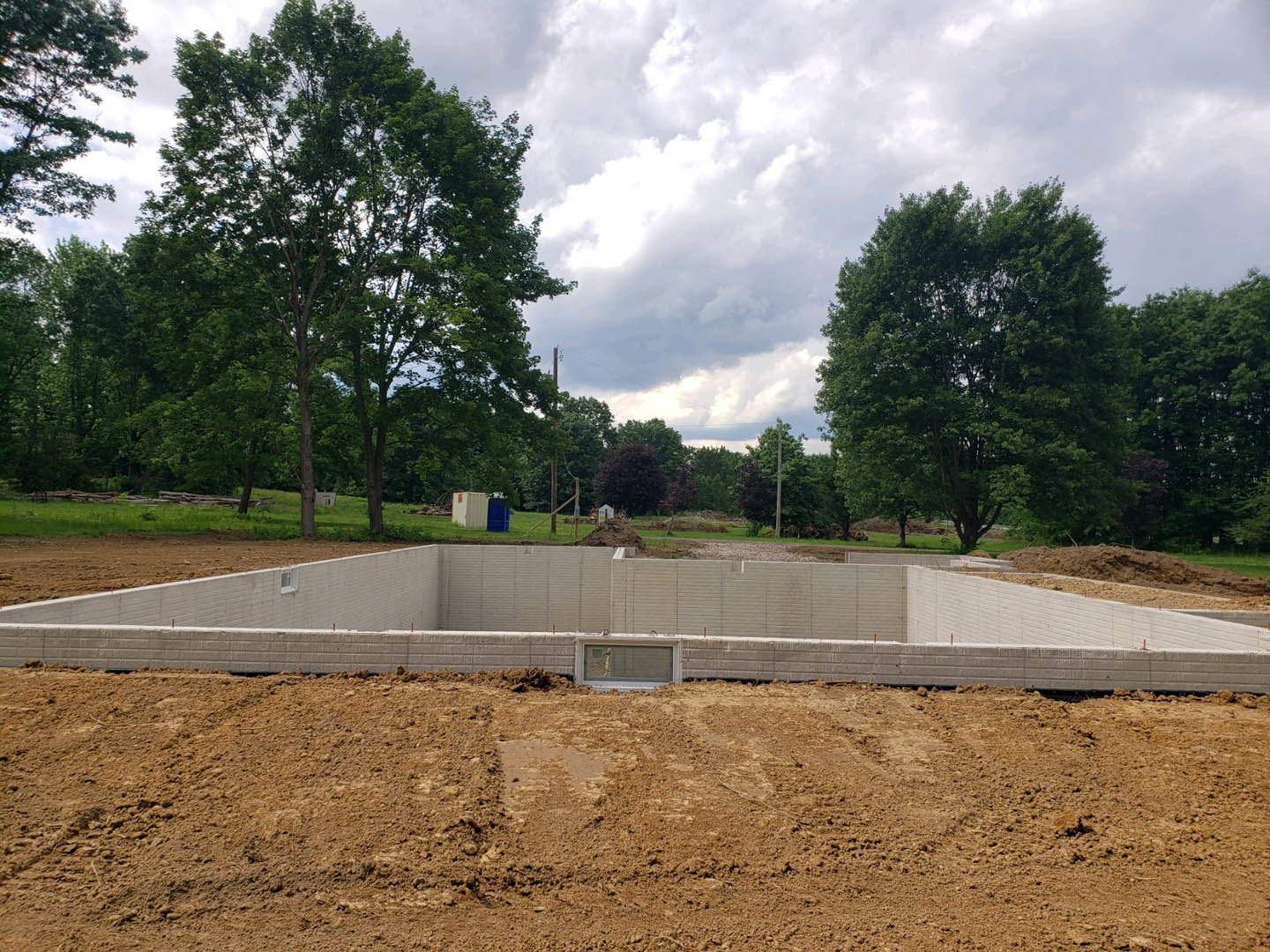 Dirt construction site bordered by mature trees under a cloudy sky, unfinished exterior wall visible in foreground