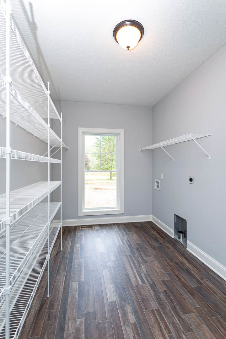 White metal shelving unit with grated shelves against a plaster wall, large window overlooking trees, hardwood floor, ceiling light fixture
