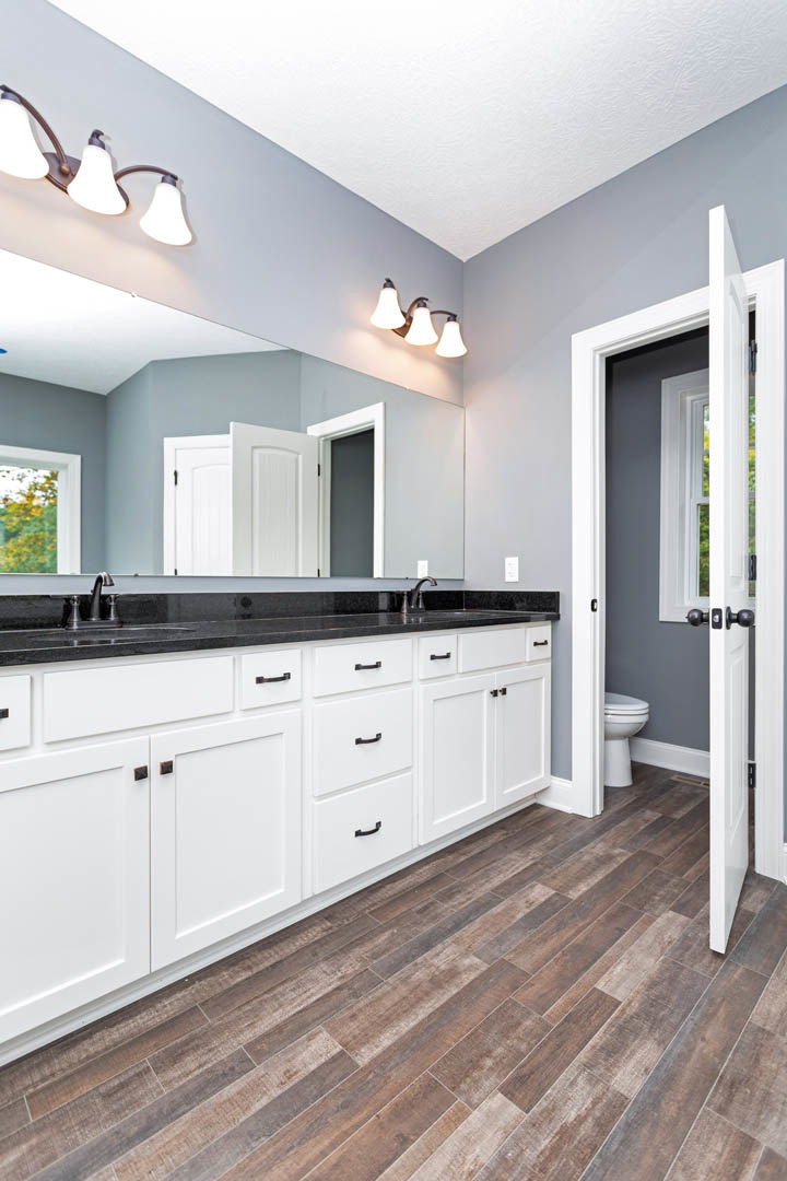 Bathroom with white cabinets, black countertops, wood flooring, three-light fixture, window showing trees, and partial view of a door
