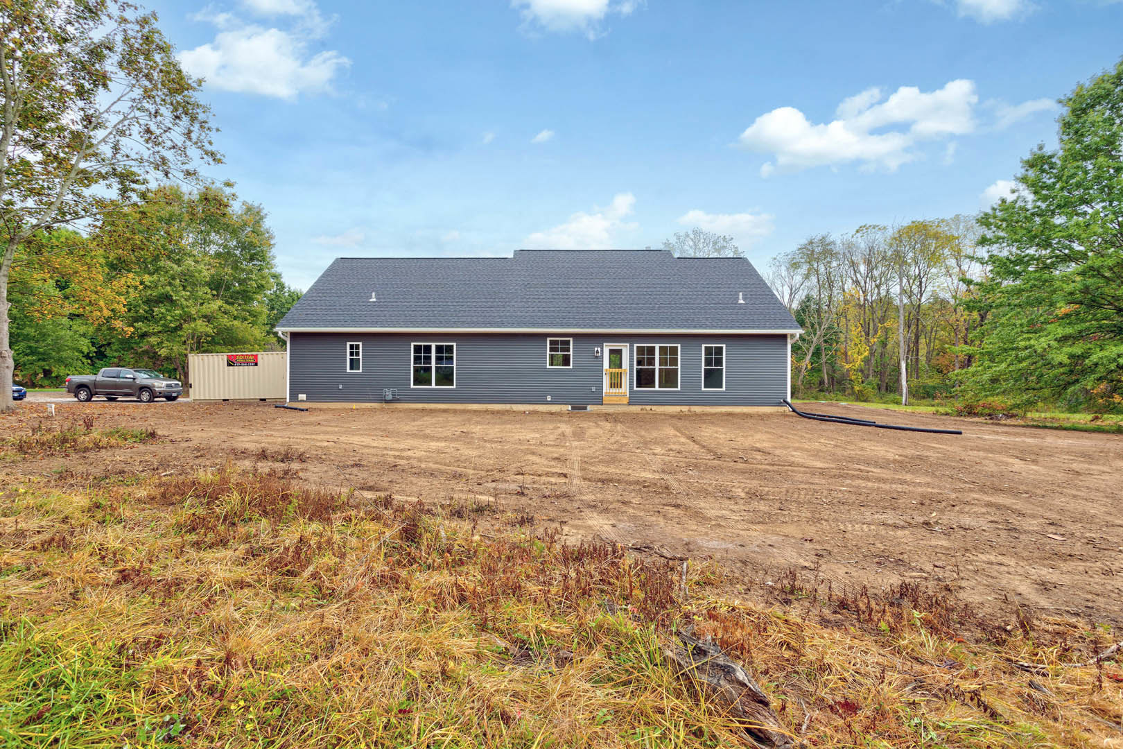 Single-story house with light siding, yellow wooden fence, dirt yard with patches of grass, parked car along roadside, metal container with sign, cluster of trees in background