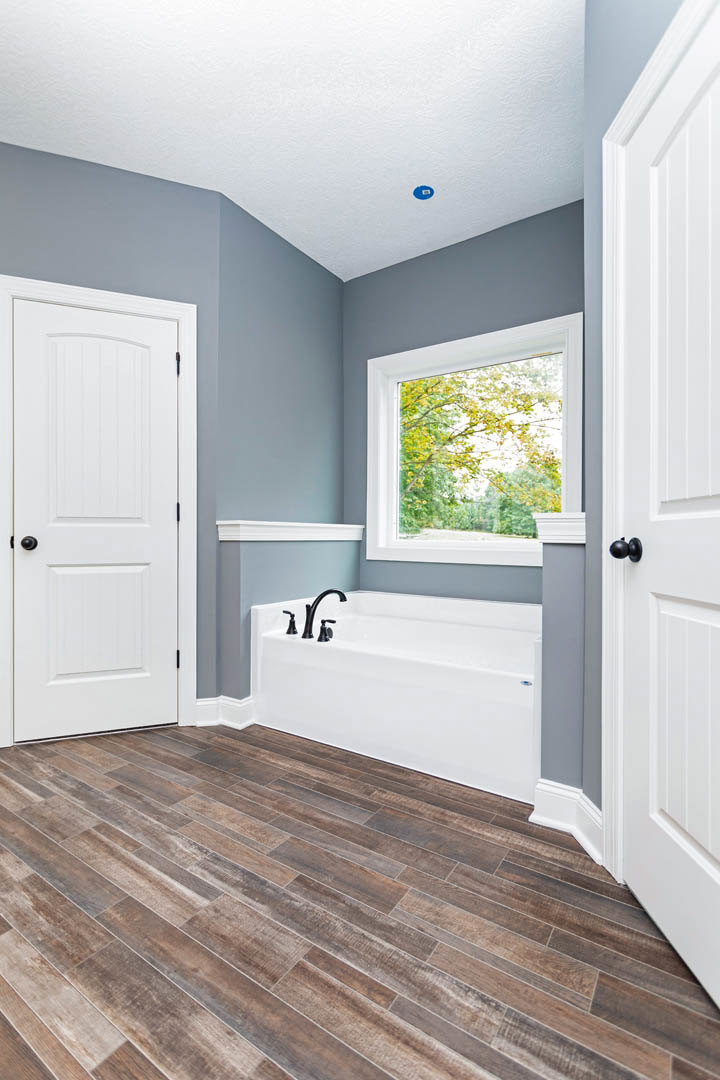 Freestanding white bathtub with chrome faucets on wood floor, large window with view of trees, white door with black knob, white trim along baseboards
