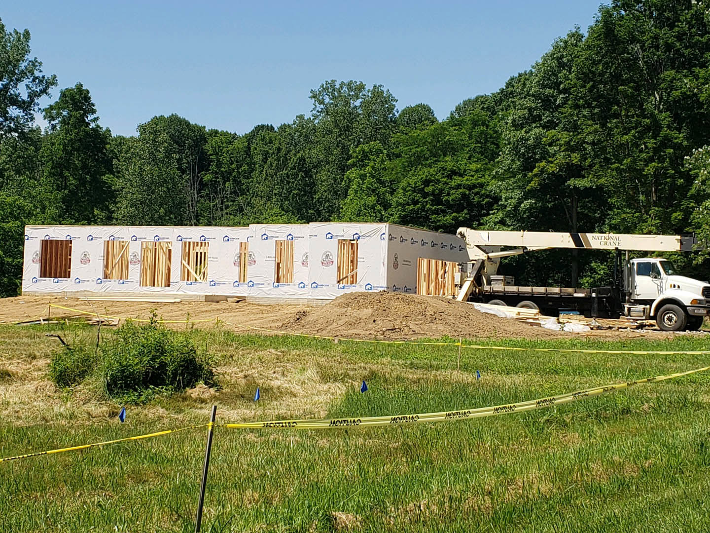 Wood-framed house under construction on grassy lot, yellow caution tape in foreground, white perforated board leaning nearby, trees and blue sky in background, crane on trailer