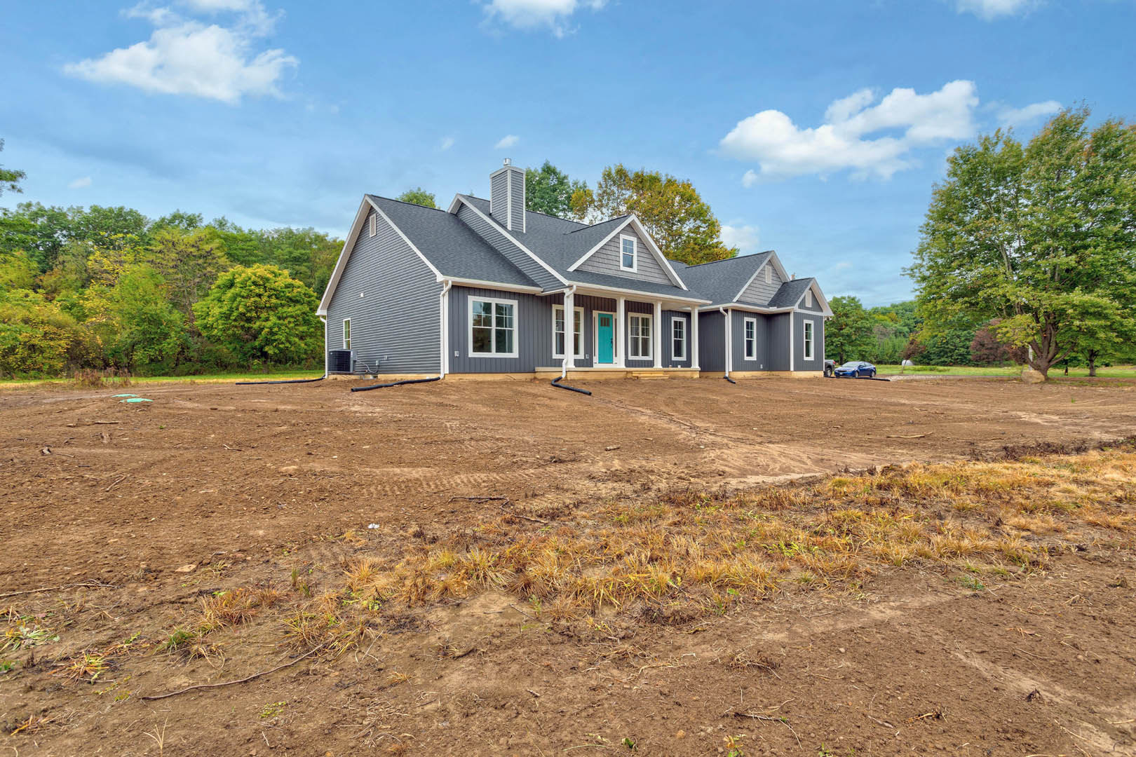 Two-story house with blue front door, white siding, and large windows, set on a dirt lot with scattered grass and pipes, mature leafy trees in the background under a cloudy sky