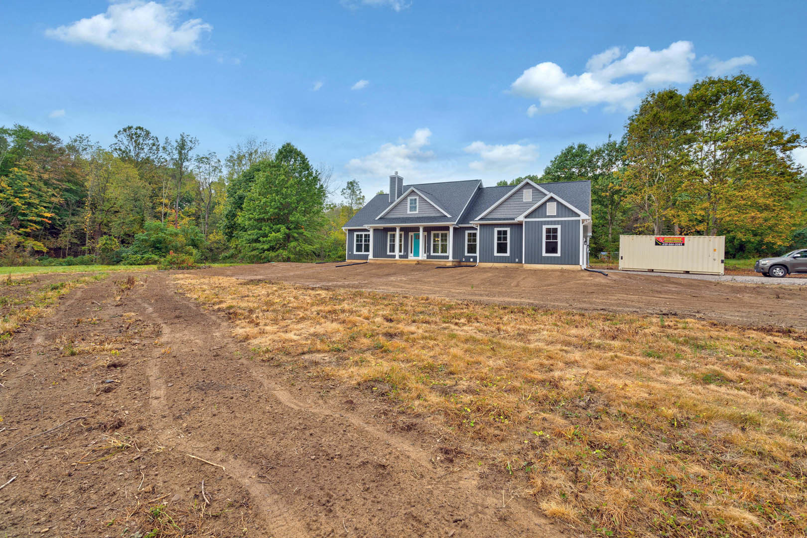 Modern home with blue front door, white exterior, dirt driveway, grassy lot, white container with signage, parked car, and blue sky with scattered clouds