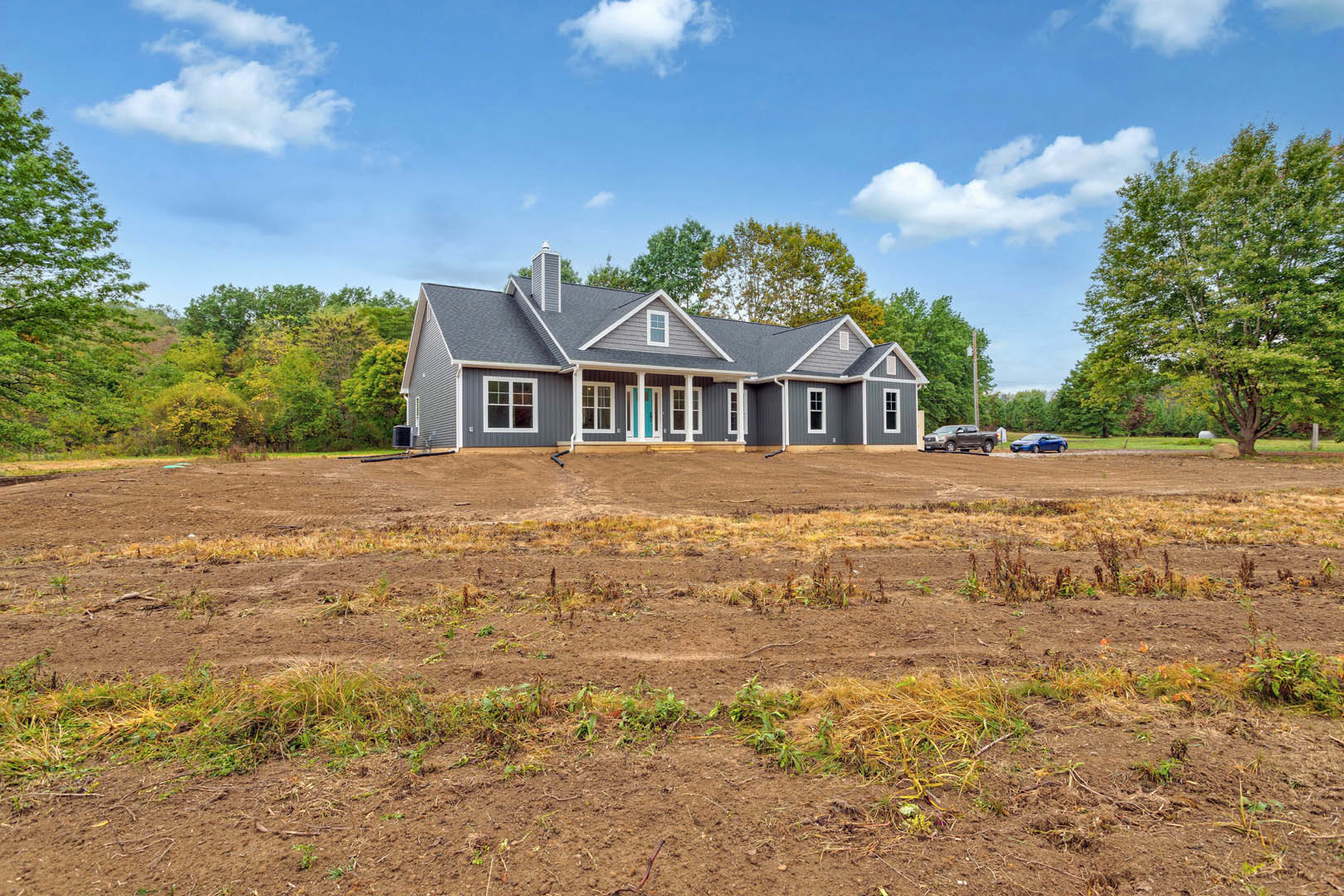White farmhouse with blue front door, surrounded by dirt field with patches of weeds, mature trees with green leaves in background, cloudy sky overhead