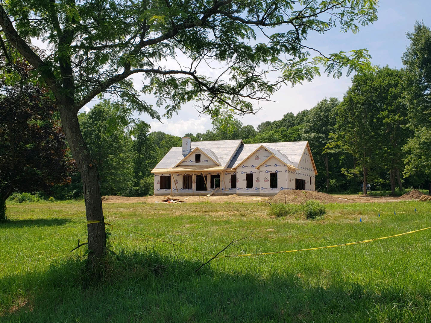 Wood-framed house under construction surrounded by tall trees and grassy field, partially enclosed by a wooden fence, blue sky overhead