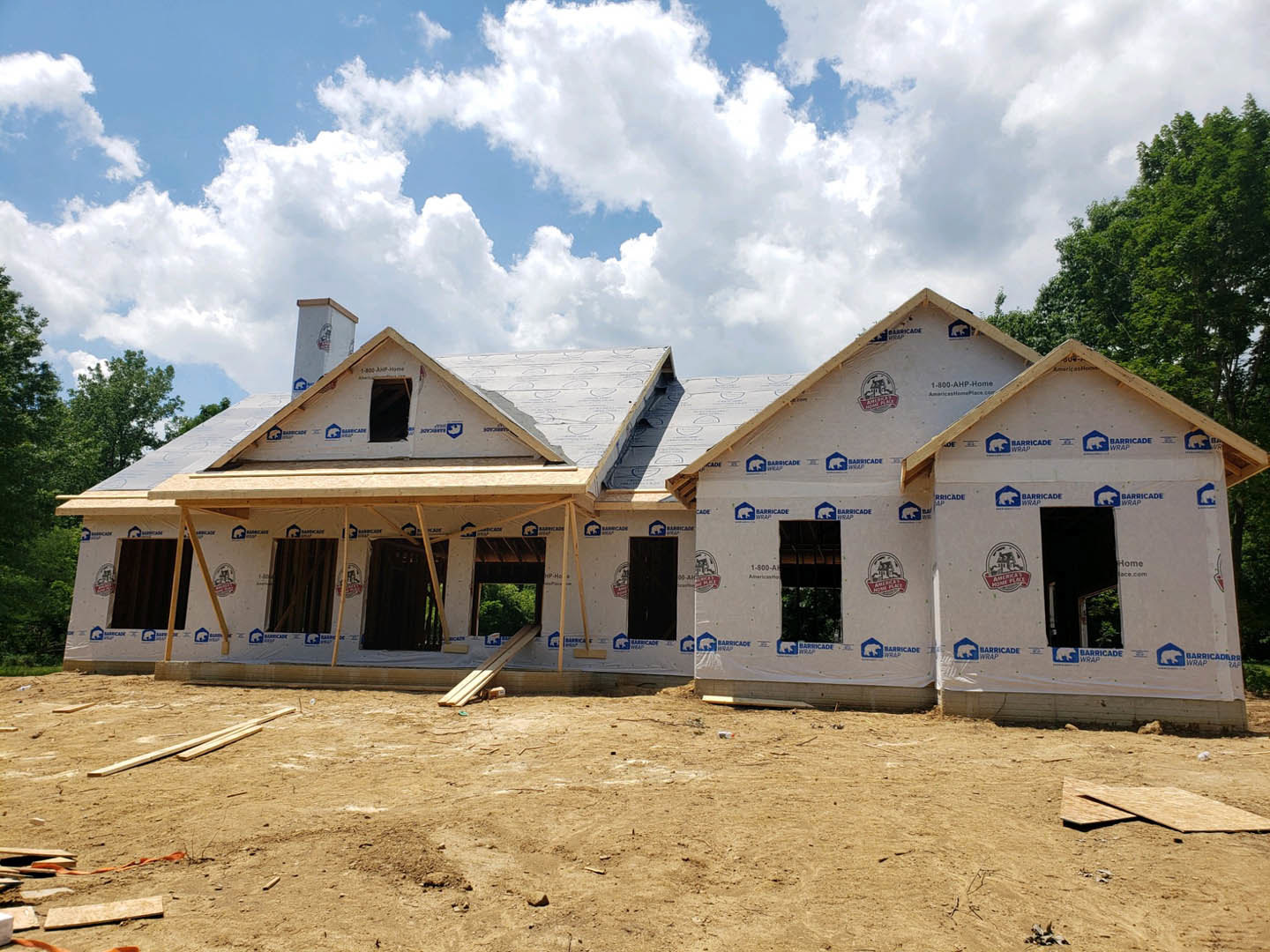 Partially built house with exposed wood beams, surrounded by several trees, cloudy sky overhead, construction materials and equipment visible, logo displayed on white wall surface.