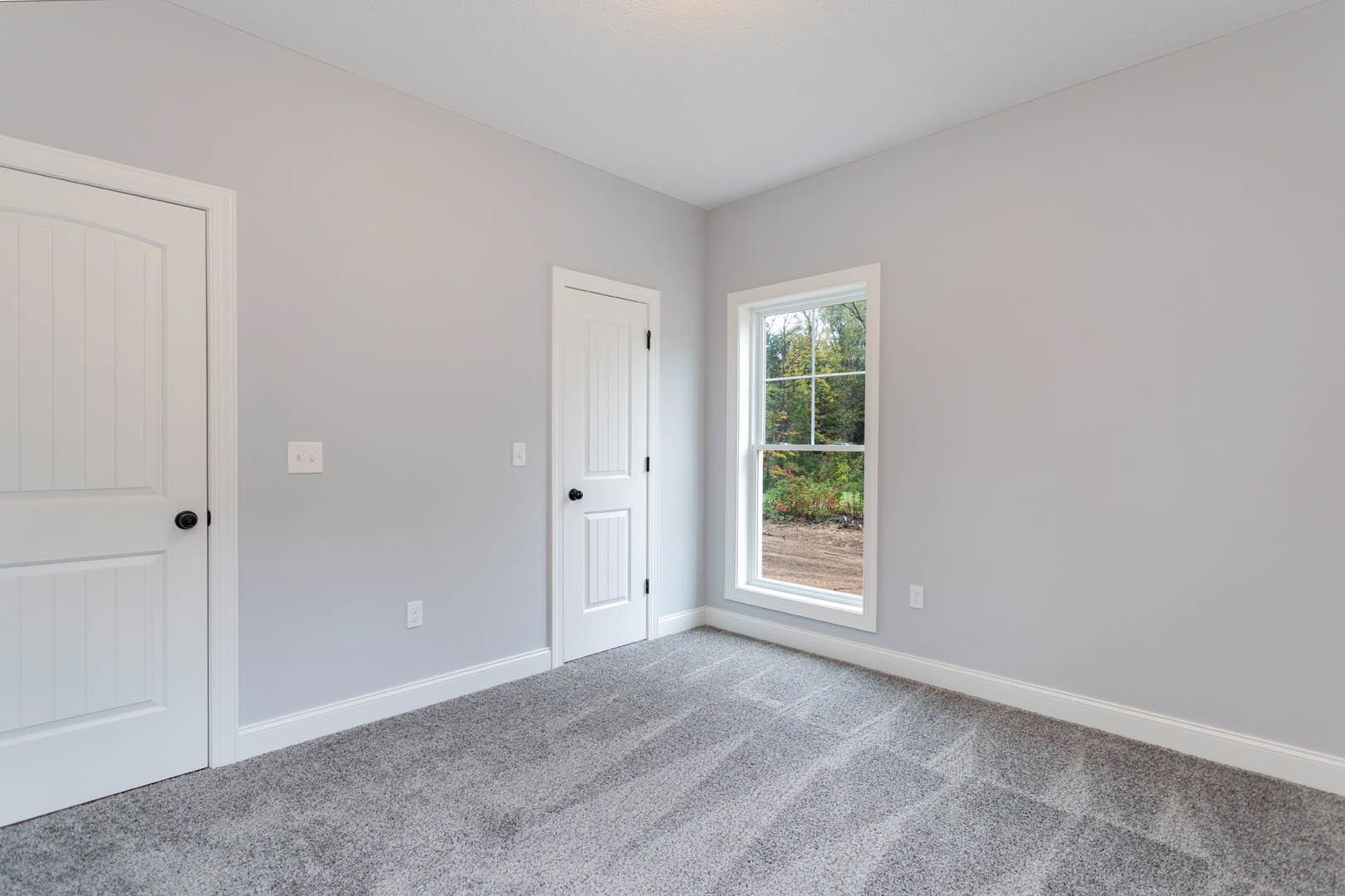 Carpeted room with white trim, white door, window overlooking forest, and white light switch with black and silver accents
