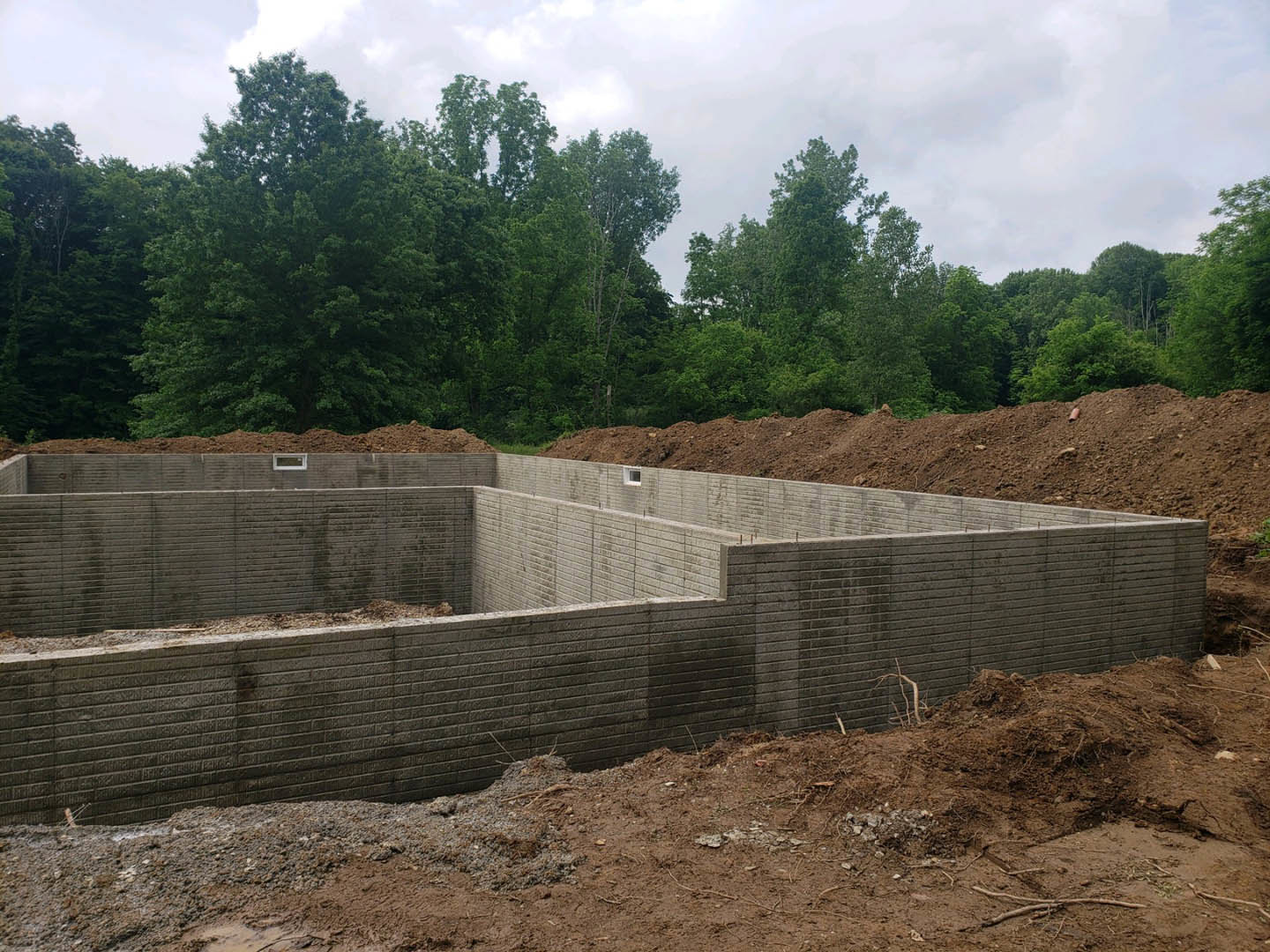 Concrete foundation with a hole in the wall, surrounded by dirt and trees, dirt road leading to site, pile of soil next to brick wall, close-up of tree branch in foreground
