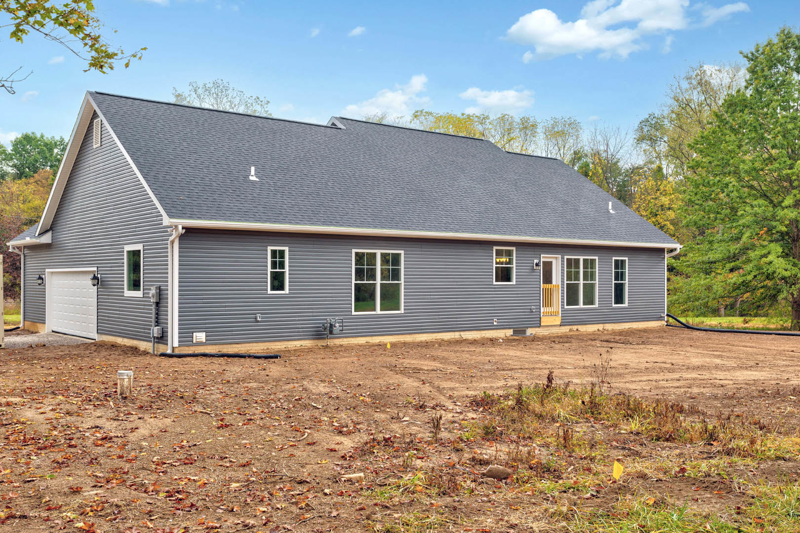 Modern farmhouse with light siding, large windows, dirt yard scattered with leaves, black metal utility box and pipes along the side, cloudy sky overhead.