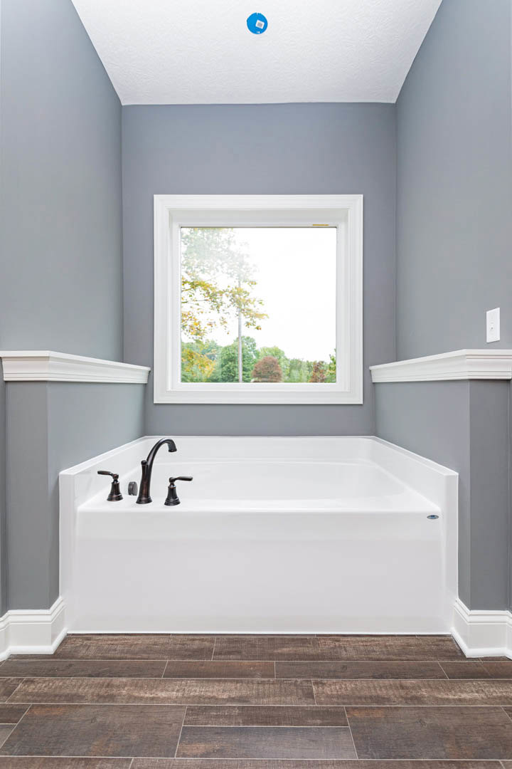 Freestanding white bathtub with chrome faucet on wood floor, large window overlooking trees, blue circular rug with white squares, light-colored walls