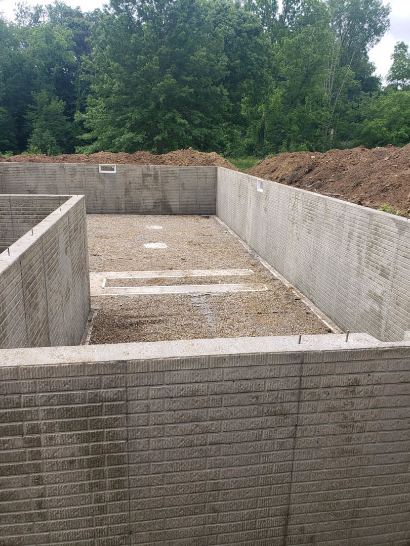 Concrete foundation slab surrounded by dirt, trees, and woodland, with a wall section featuring a window opening and a pile of soil nearby.
