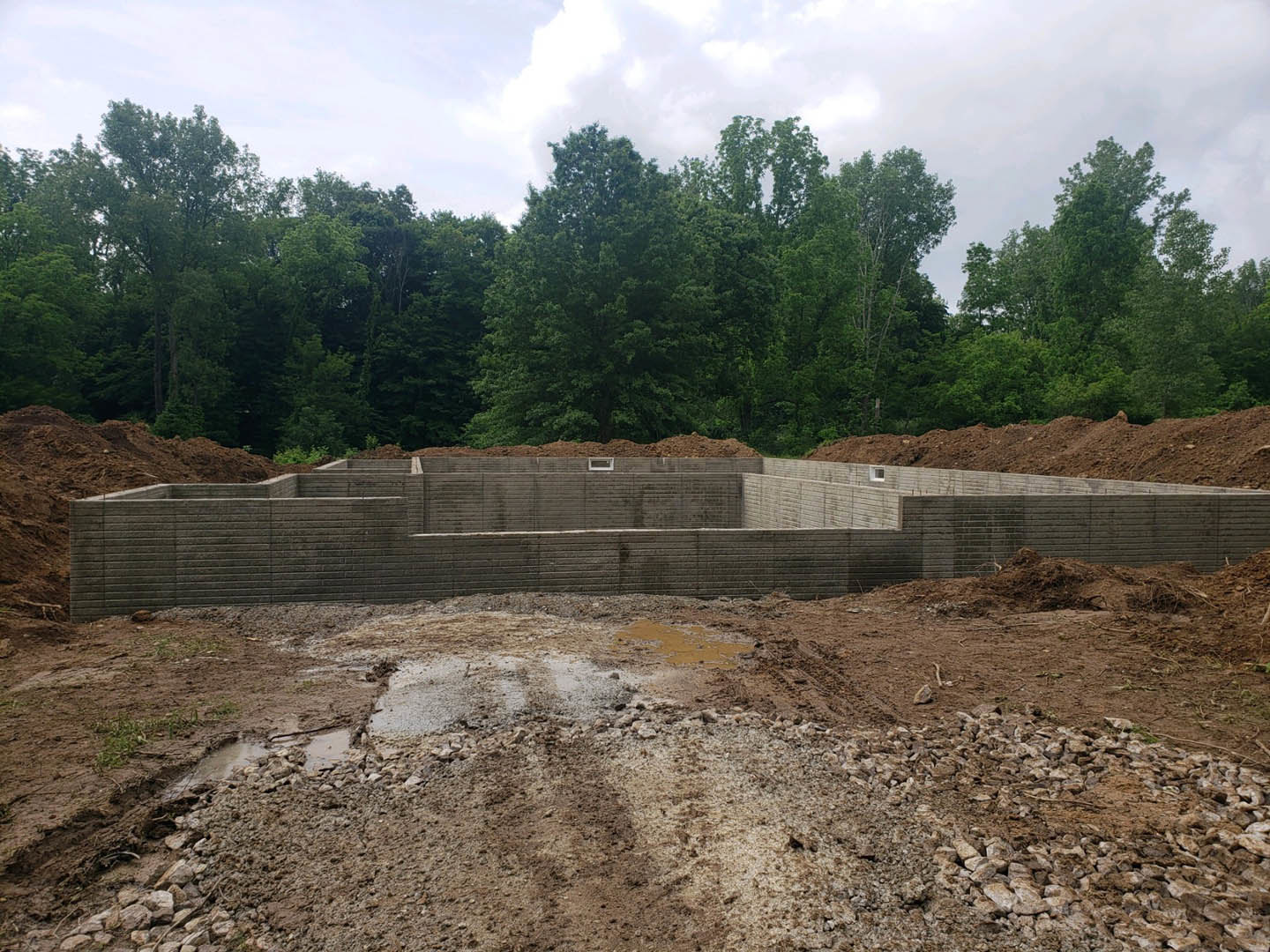 Concrete foundation slab surrounded by dirt, rocks, and muddy ground, bordered by trees under a cloudy sky