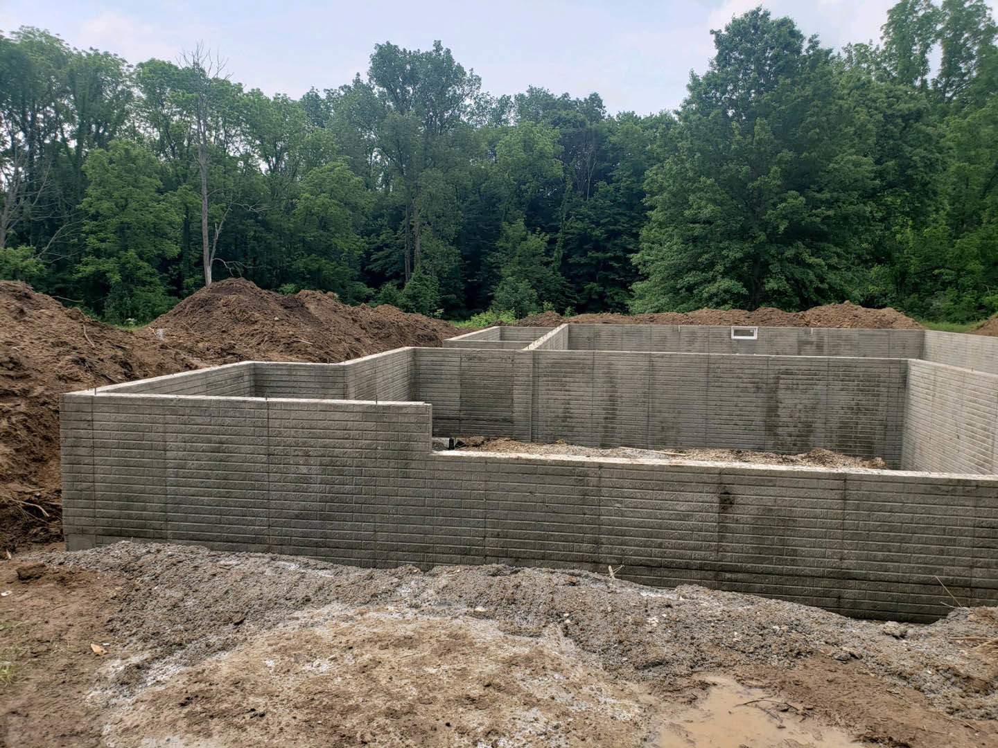 Concrete foundation and partial brick wall surrounded by piles of dirt, with mature trees and blue sky in the background