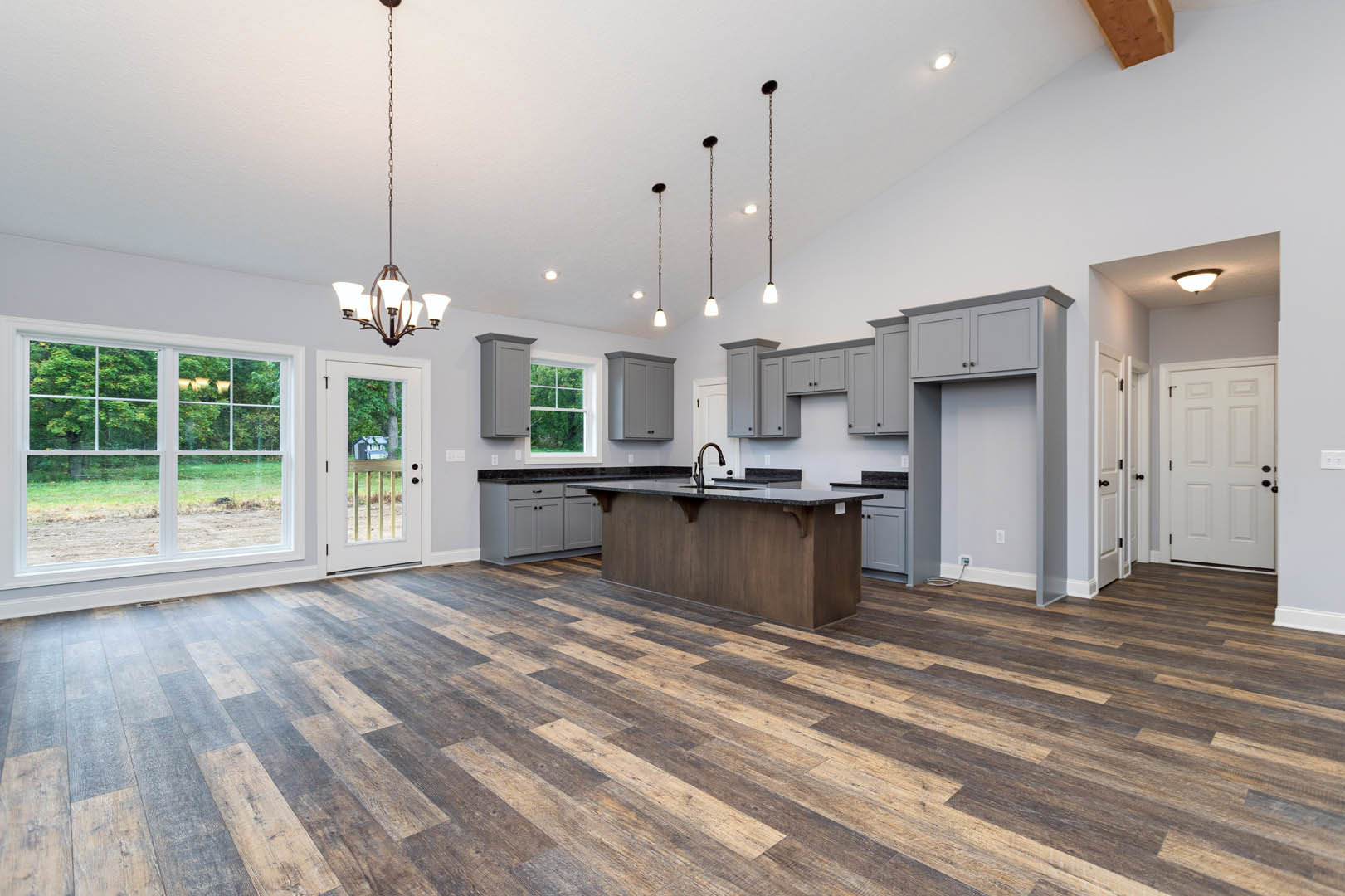 Spacious kitchen featuring wood flooring, white cabinetry with black knobs, a close-up of a countertop, black faucet, and a window overlooking a field of trees.