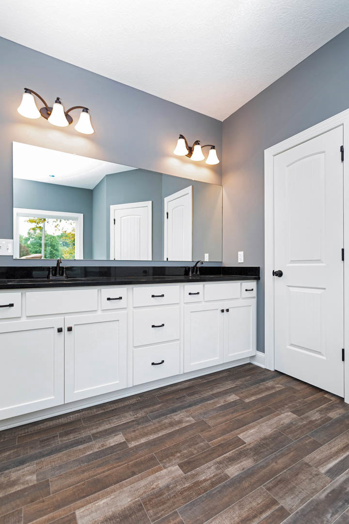 Bathroom with white cabinets, black countertops, wood flooring, white door with black knobs, window showing trees, and three-light fixture