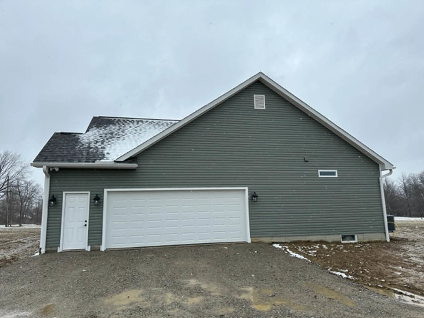 White garage door with black handles, gray shingle roof, light siding, small white wall vent, dirt driveway, partly cloudy sky