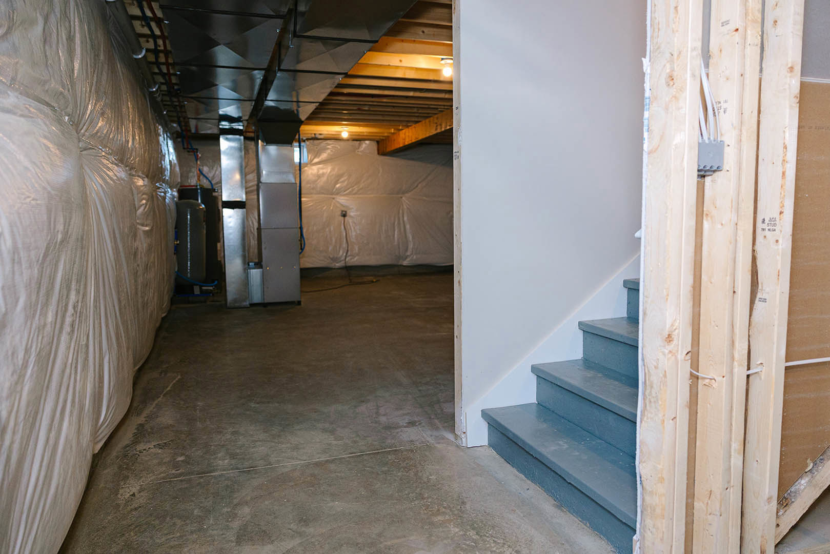 Basement with unfinished concrete floor, staircase with wooden steps, wall covered in clear plastic sheeting, exposed metal ceiling, visible building insulation.