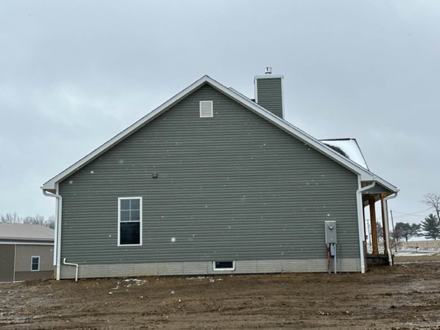 Gray siding house with brick chimney, covered front porch, white-framed windows, white wall vent, dirt yard, and cloudy sky