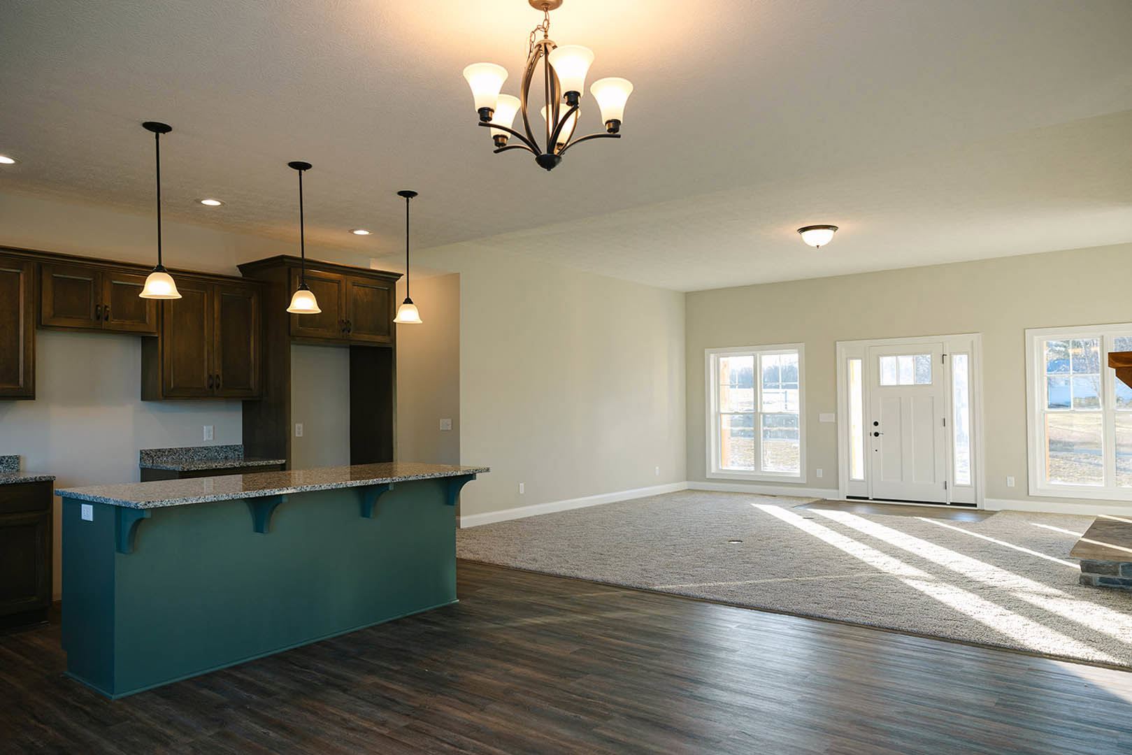 Open-concept living room with wood flooring, kitchen island featuring a countertop, blue accent wall, five-light chandelier, white door with glass panes, and a window close-up