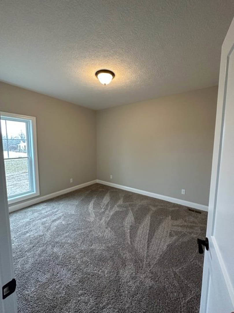 Carpeted room with white walls, ceiling-mounted light fixture, large window overlooking grassy field, black rectangular vent near baseboard.