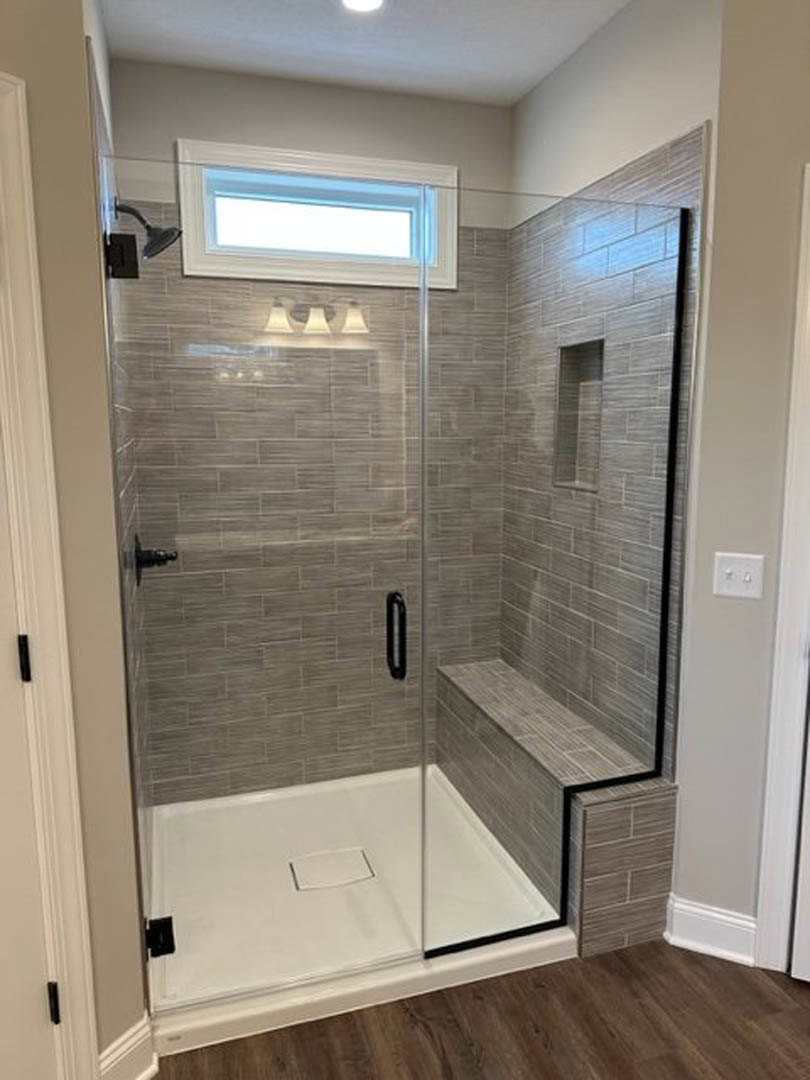 Glass shower enclosure with built-in bench, wood floor with white trim, white wall outlet and switches, close-up of light fixture, white wall with linear detail.