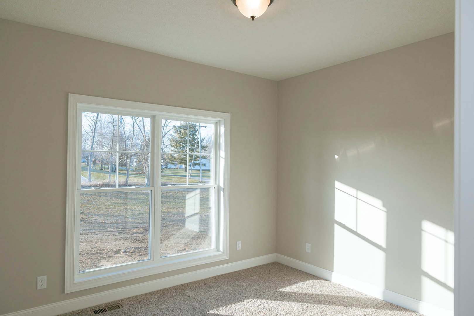 Spacious room featuring a large window with views of a grassy field and trees, white plaster walls, crown molding, neutral carpet flooring, and a modern light fixture.