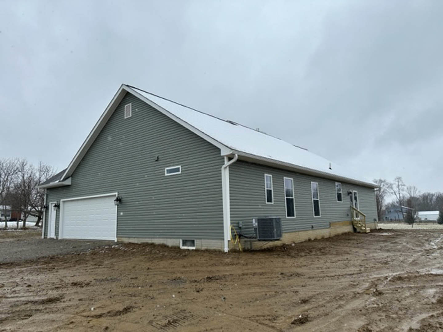 Two-story house with white siding, attached garage with white-framed door, snow covering roof, dirt yard in foreground, overcast sky