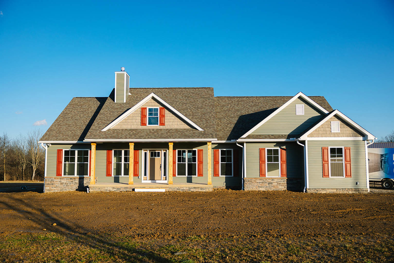 Two-story farmhouse with white siding, front porch, dark shutters, dormer window, brick chimney, and grassy yard in the background