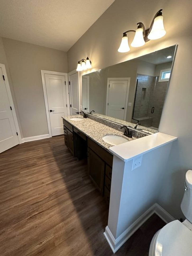 Bathroom with double sinks set in a white marble countertop, large wall-mounted mirror above, wood flooring, three-light fixture, small square outlet, and visible toilet paper