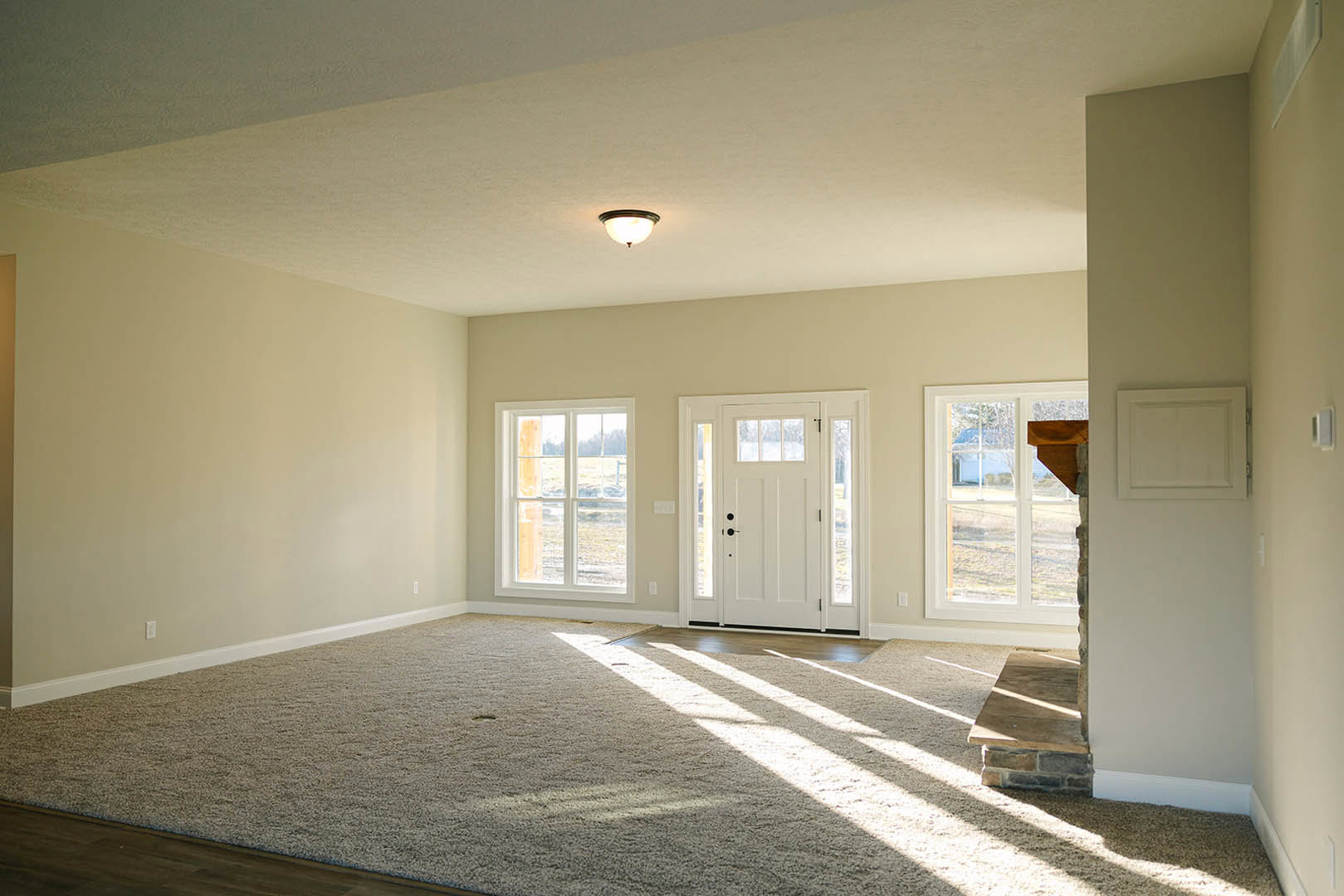 Carpeted living room with stone fireplace, white framed windows, white door with glass panels, round ceiling light fixture, sunlight streaming onto floor