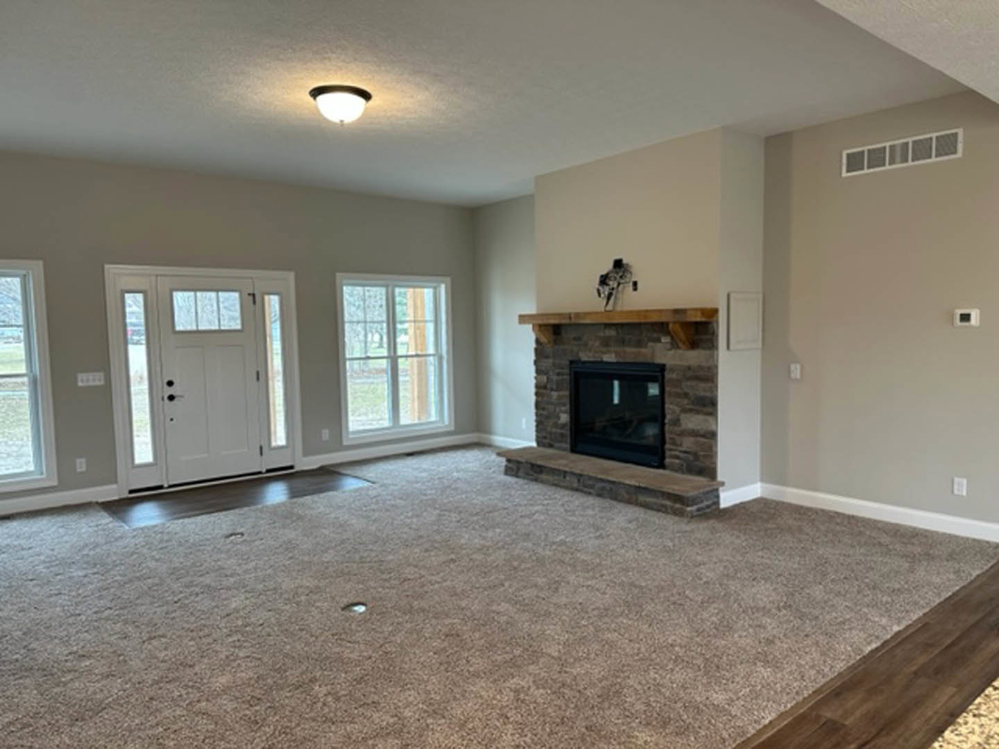 Carpeted living room featuring a central fireplace with a wooden mantel, white door with glass panes, wall vent, and neutral walls and ceiling.