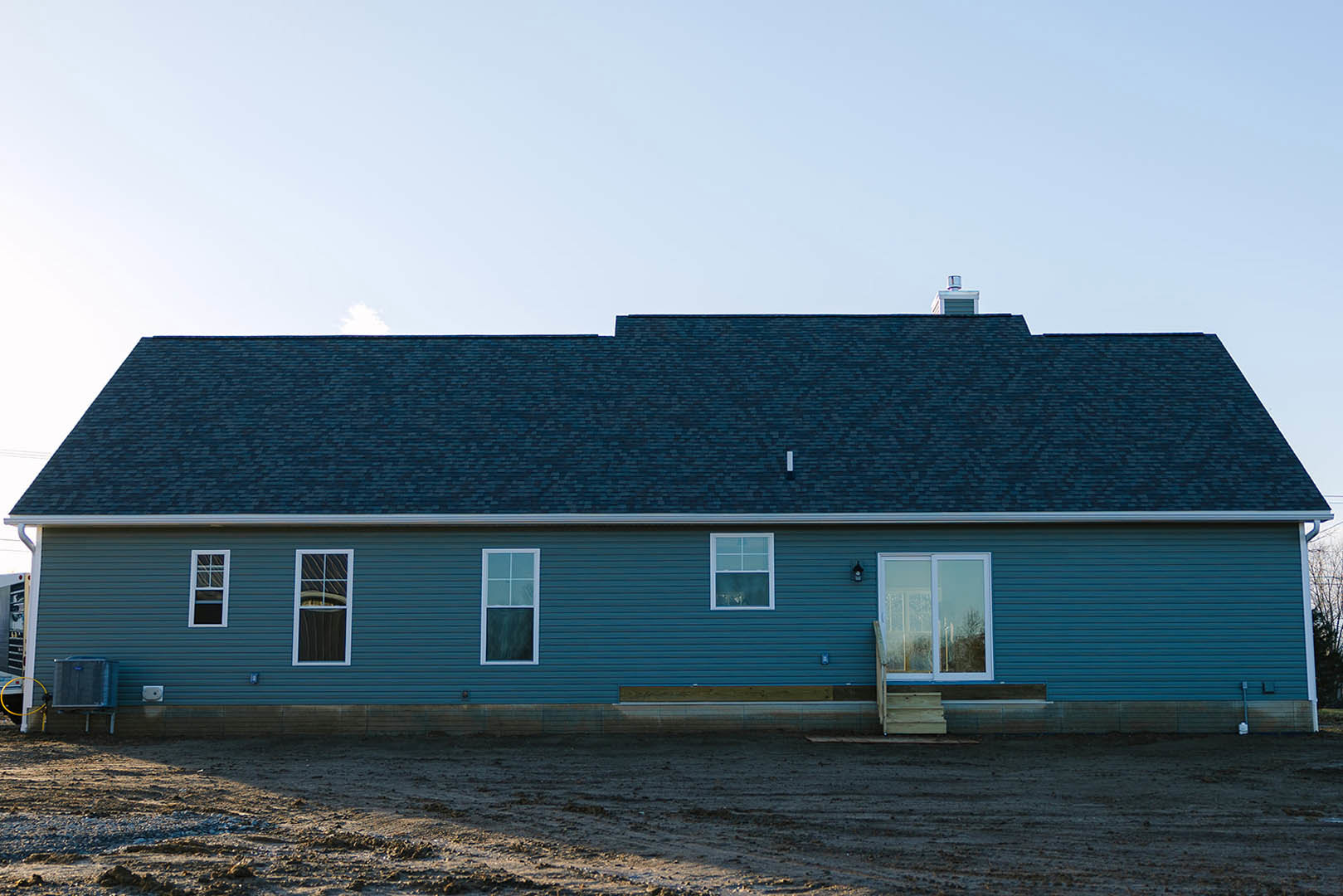 Single-story house with blue metal roof, white-framed windows, sliding glass door, and light-colored siding, set beside a dirt field under a clear sky