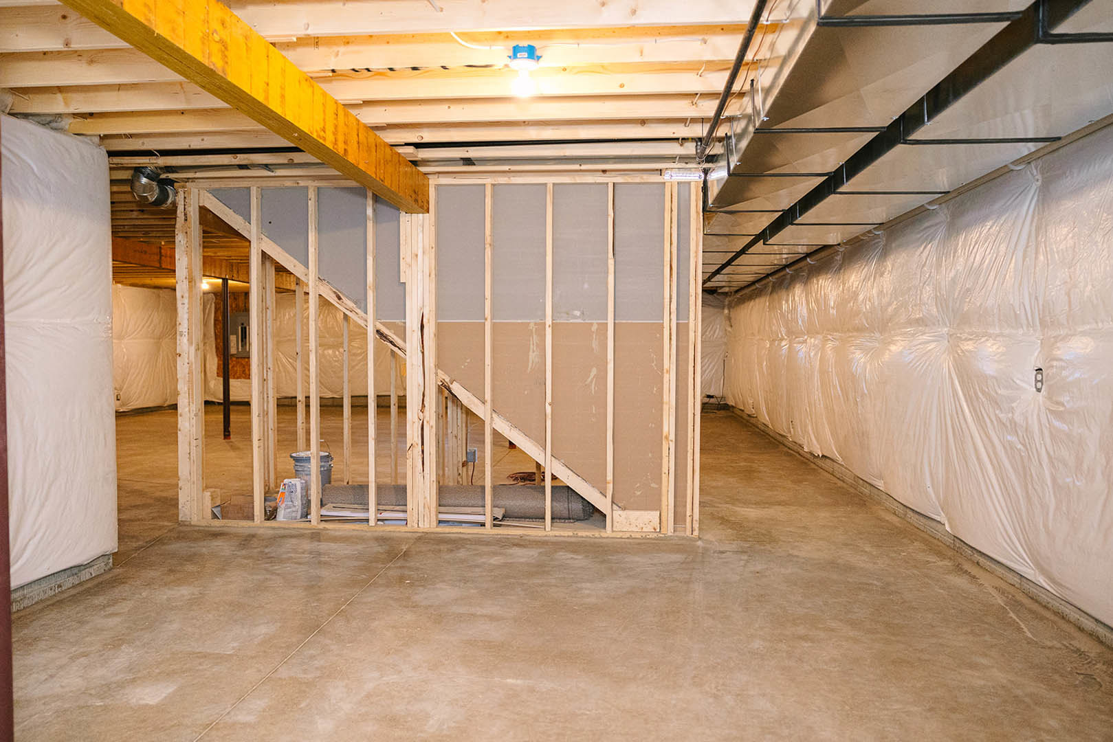 Basement room with exposed metal beams, plastic wall covering, plaster walls, white sheet draped over bed, staircase centered against wall, blurry blue object in foreground.