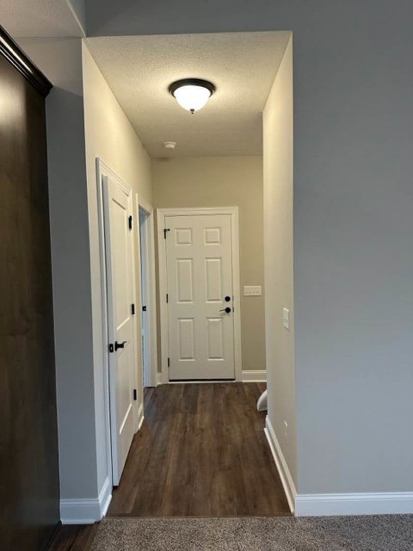 Hallway with white paneled doors featuring black knobs, hardwood flooring with white baseboard trim, ceiling-mounted light fixture, and adjacent wood wall and carpeted area