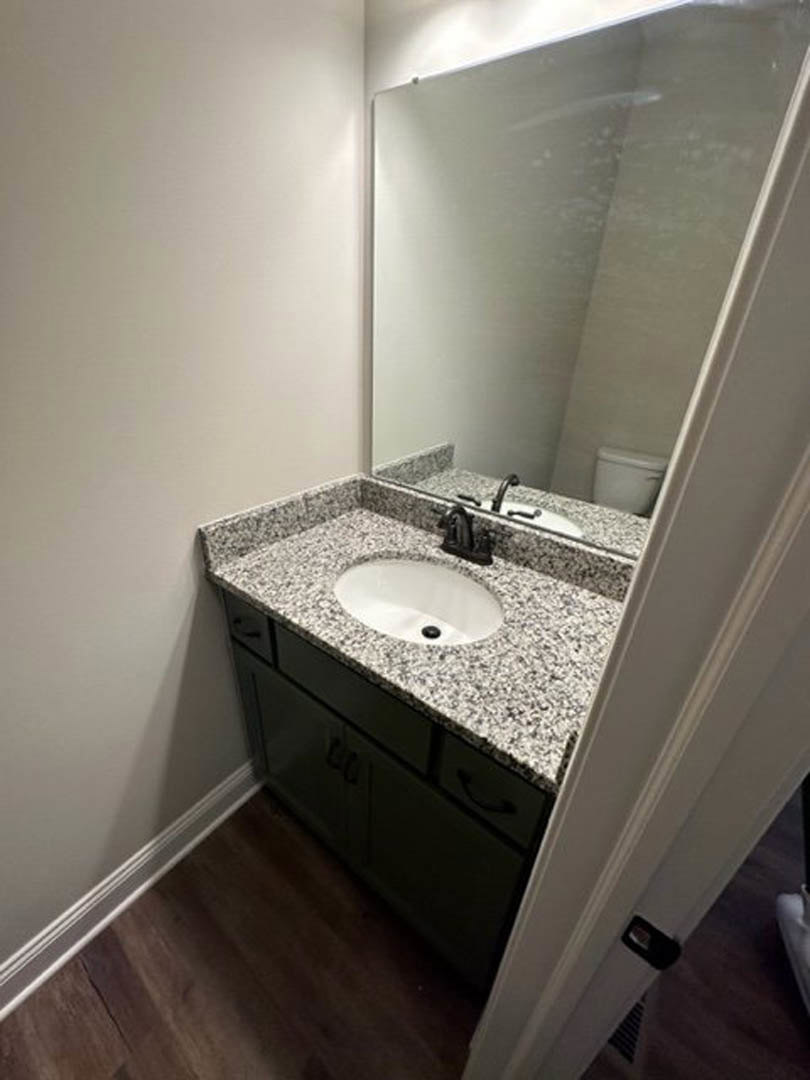 White porcelain sink with chrome faucet set in a stone countertop, rectangular wall mirror above, light gray tile backsplash and floor, modern bathroom accessories visible