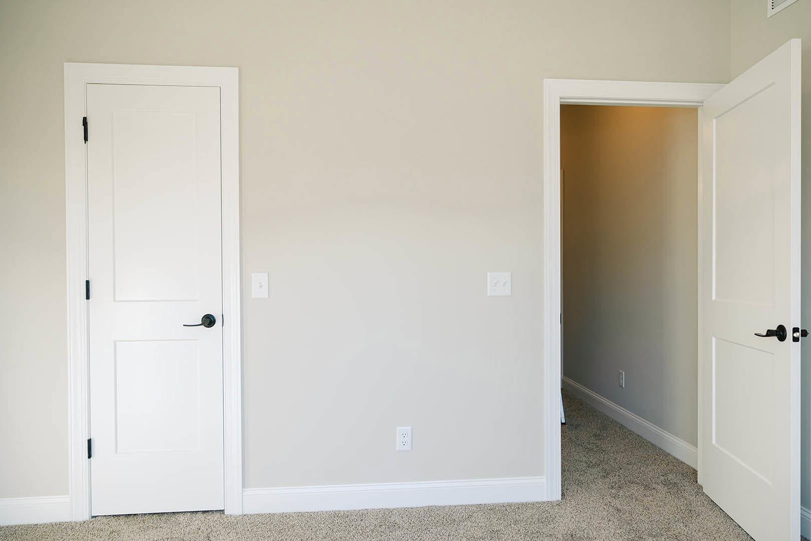 White paneled door with black handle, open to reveal adjoining room; white walls, white trim, and dual wall switch visible; light wood flooring throughout.