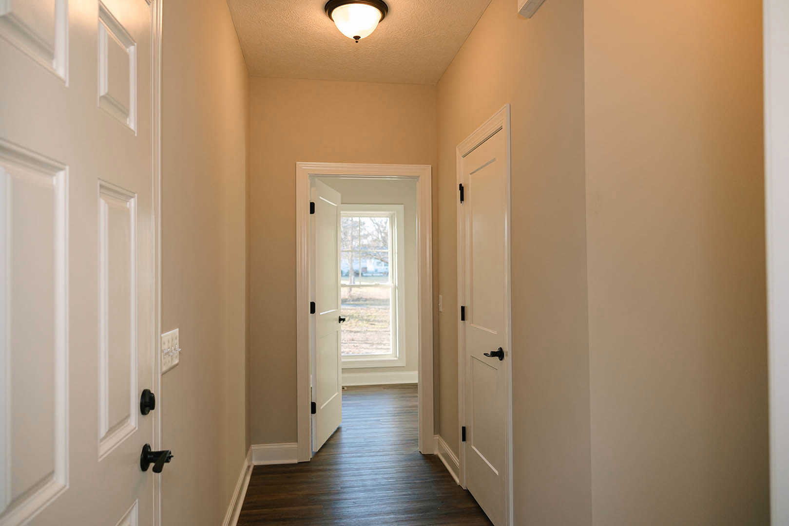Hallway with wood flooring, white paneled doors featuring black handles, round ceiling light fixture, white walls with molding, and a light switch near the doorway.