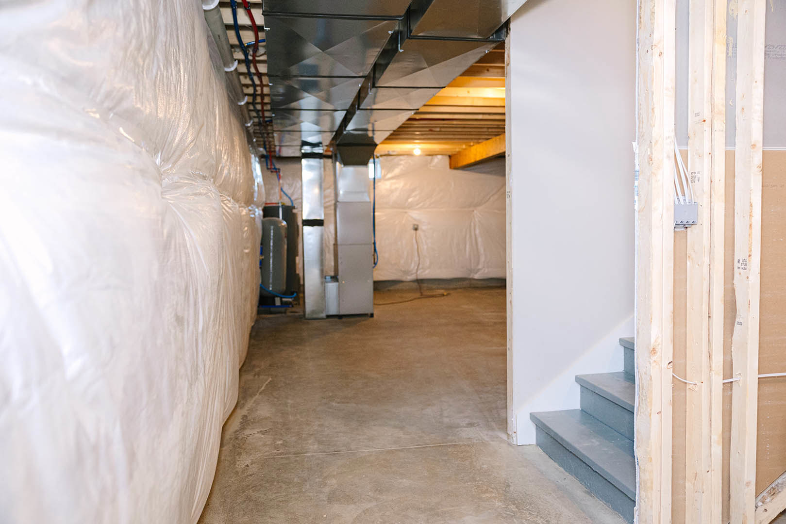 Basement with smooth white plaster wall, exposed metal pipes along ceiling, concrete floor, and white plastic sheeting covering part of the wall