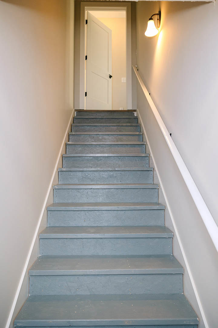 Wood staircase with black metal handrail, white plaster walls, light fixture mounted above, white door with black handles in the background, blue accent surface and floating shelf
