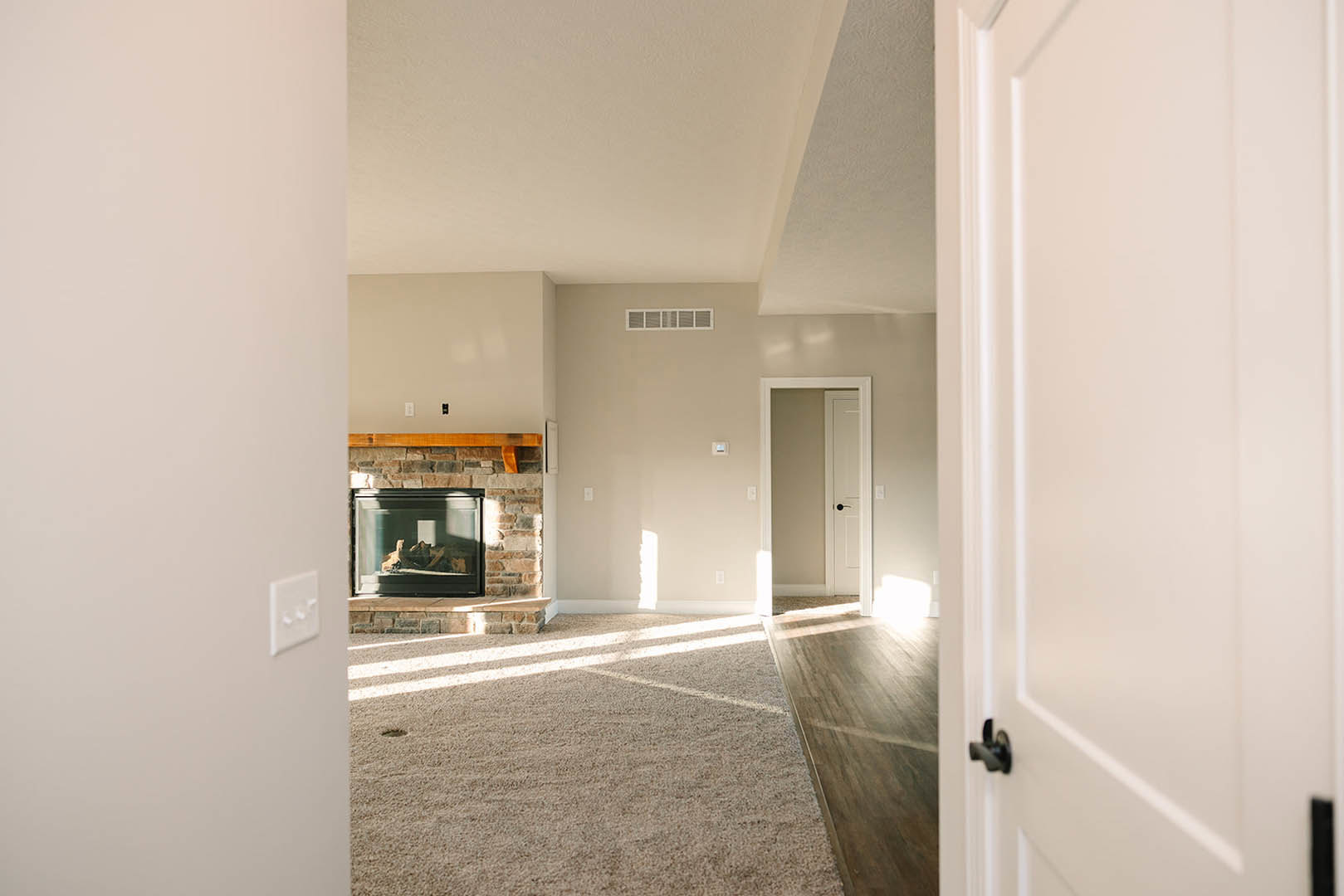 Living room with beige carpet, white fireplace with glass door, large window, and white door featuring a black handle