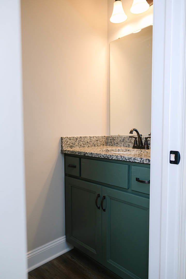 Granite bathroom countertop with undermount sink, green cabinetry, wall-mounted mirror, chrome faucet, and light fixture against green tile wall