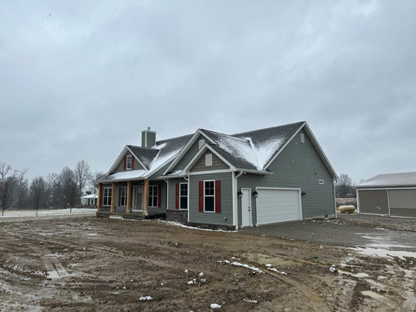 Two-story house with snow-covered roof, white-framed windows, open front door, dirt road and patch in front, man standing near garage door, overcast sky and trees in background