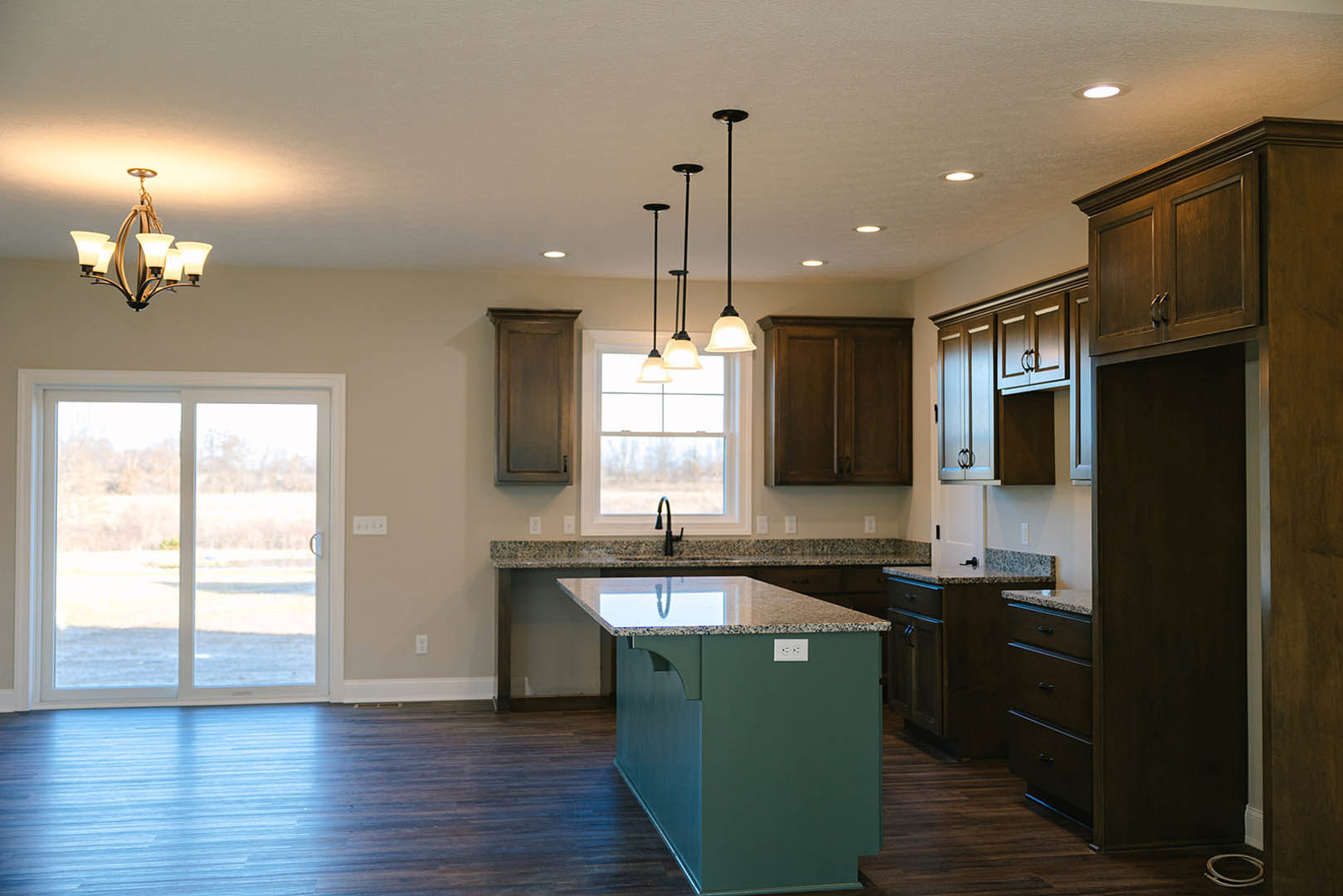 Kitchen with hardwood flooring, central island featuring granite countertops, white cabinetry, stainless steel sink, five-light fixture above, and large window overlooking outdoor
