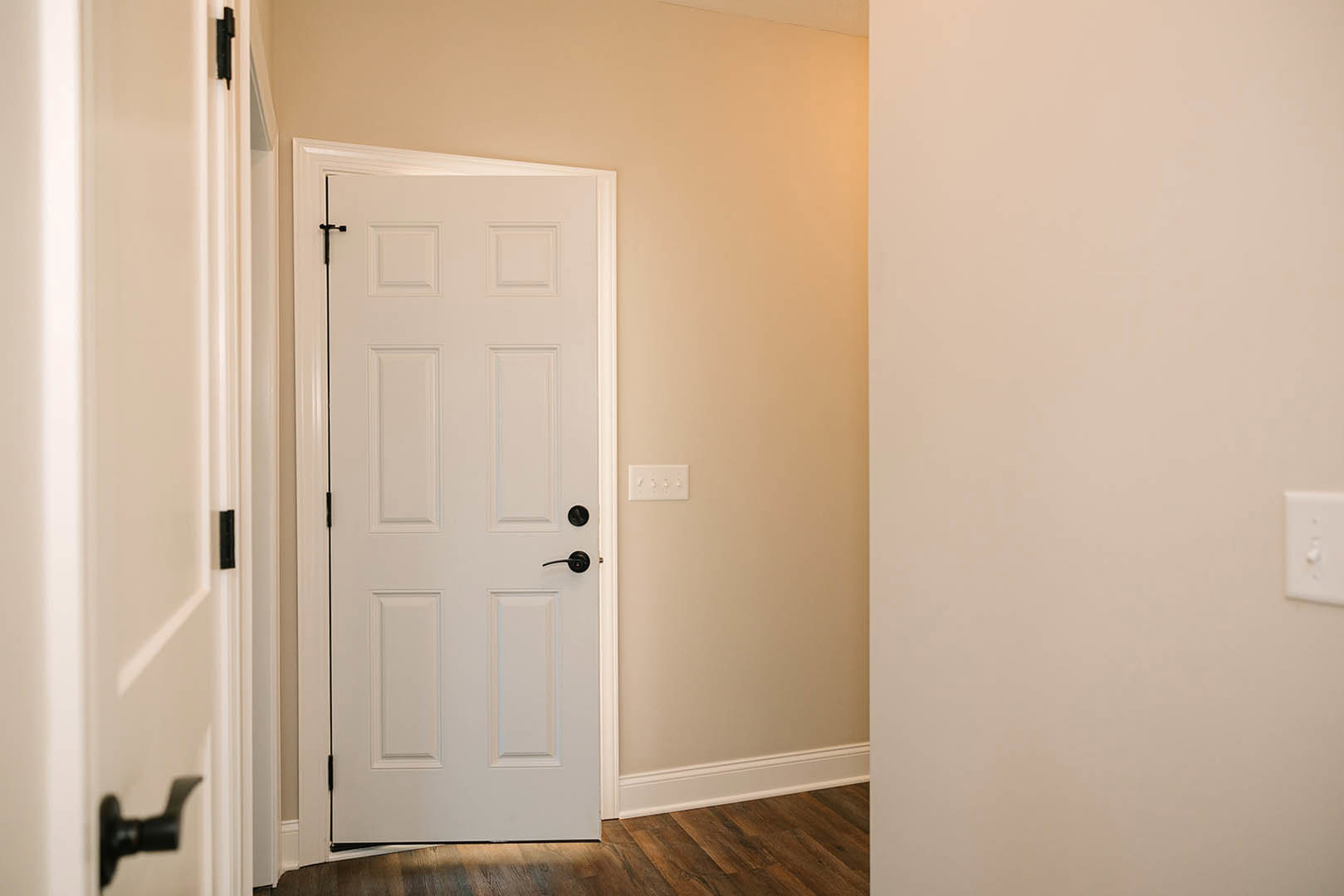 White paneled door with black handle, white electrical outlet and light switch on adjacent wall, light-colored flooring