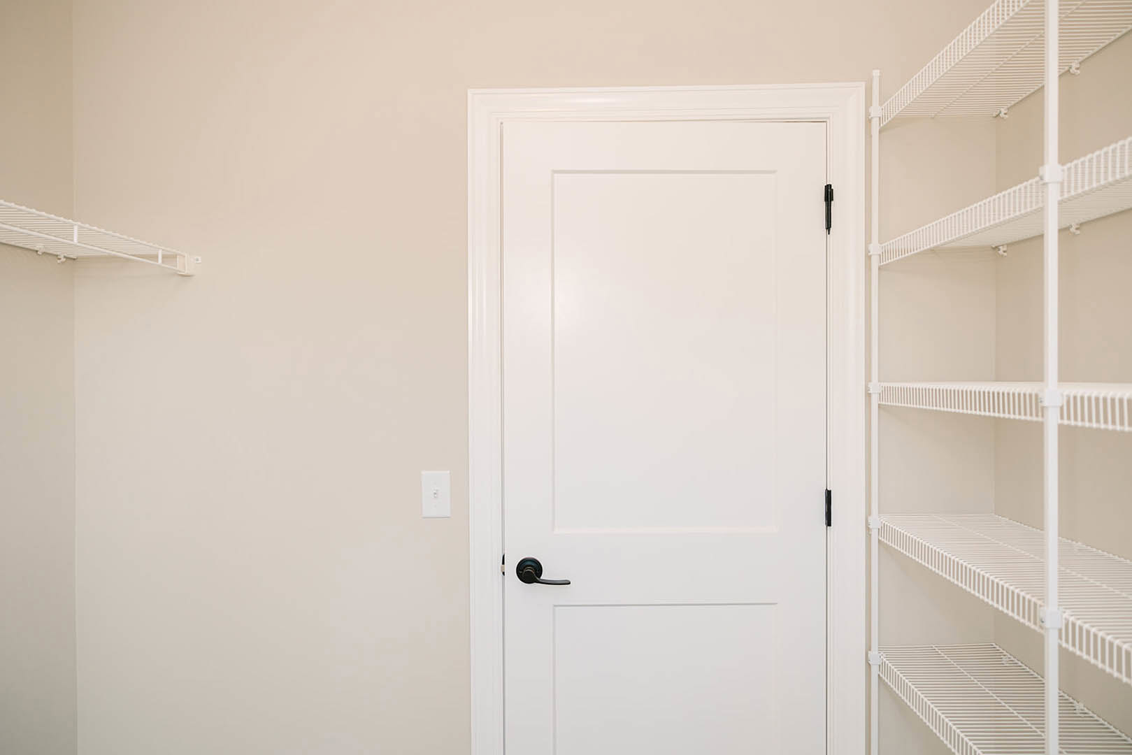 White door with matte black handle, adjacent white wall-mounted shelf, light switch visible on wall, minimalist interior design